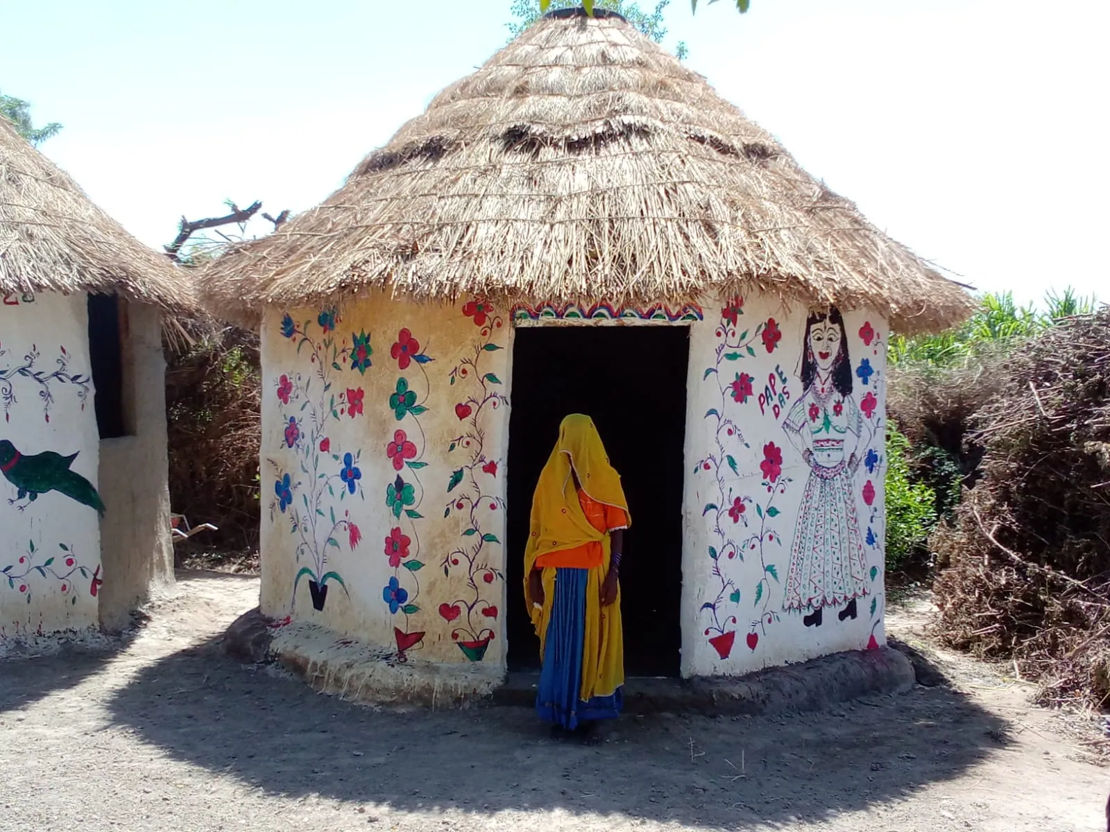 Two round homes with thatched roofs and hand-painted flowers on their walls, a woman in a yellow scarf at a doorway.