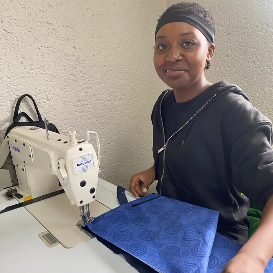 Ashley seated at an industrial sewing machine with blue fabric, smiling at the camera.